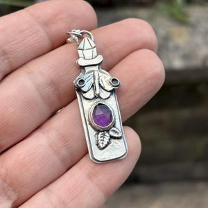 May include: A silver pendant shaped like a bottle with a faceted purple gemstone set in the centre. The pendant features a decorative moth design at the top and small leaf details at the bottom, with a crystal-like stopper at the very top.