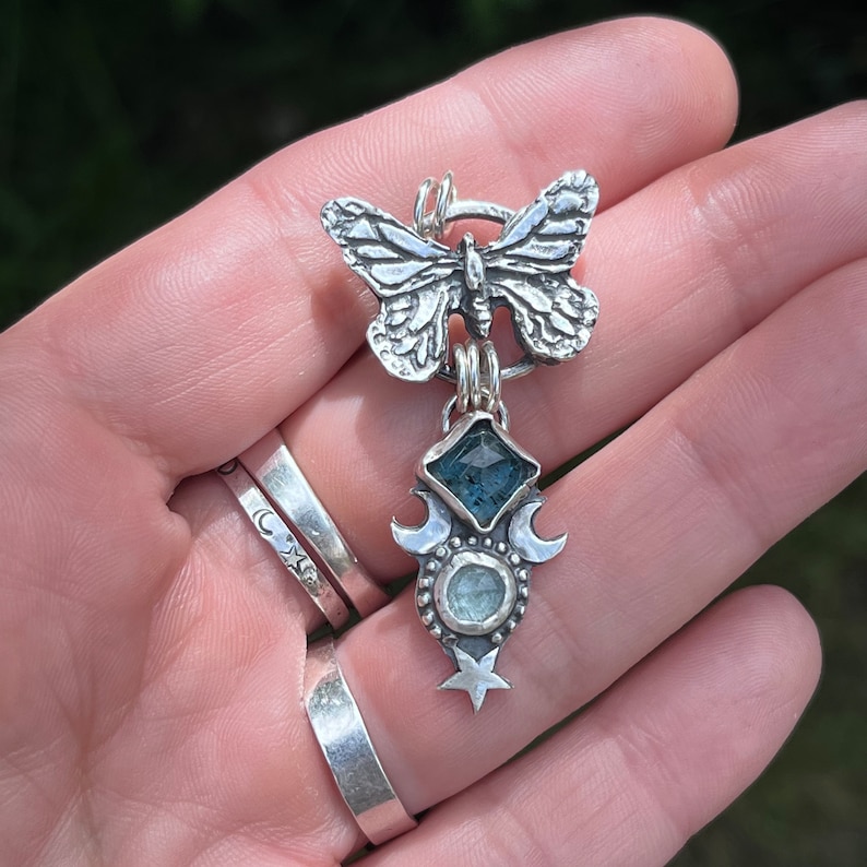 May include: A silver pendant featuring a detailed butterfly charm, a teardrop-shaped aquamarine stone, and a crescent moon design with a star.