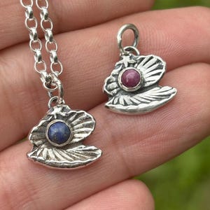 May include: Two silver pendants, one on a chain, featuring a seashell design with a central gemstone. One pendant has a blue gemstone, the other a red gemstone. The pendants are detailed with textured silver and are displayed against a blurred background.