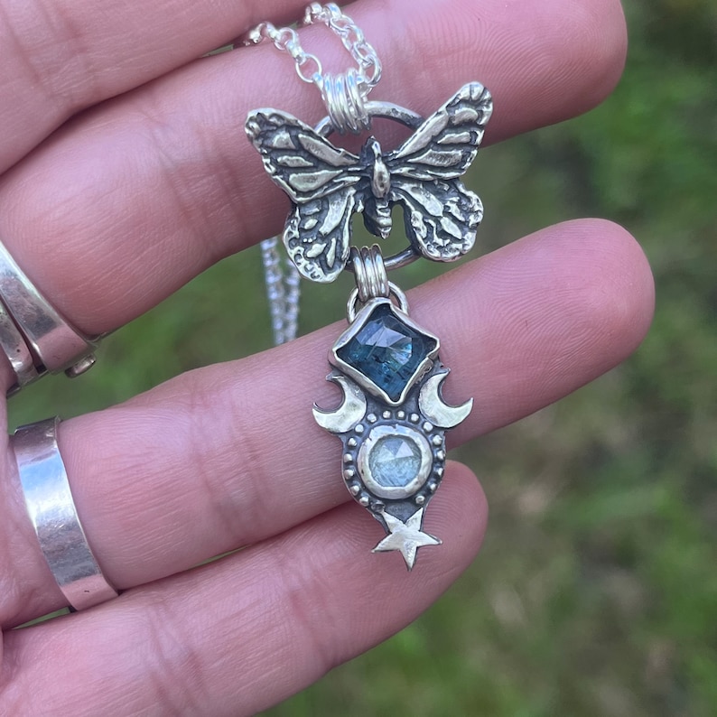 May include: A silver necklace with a butterfly pendant and a charm featuring a blue gemstone, two crescent moons, and a star.