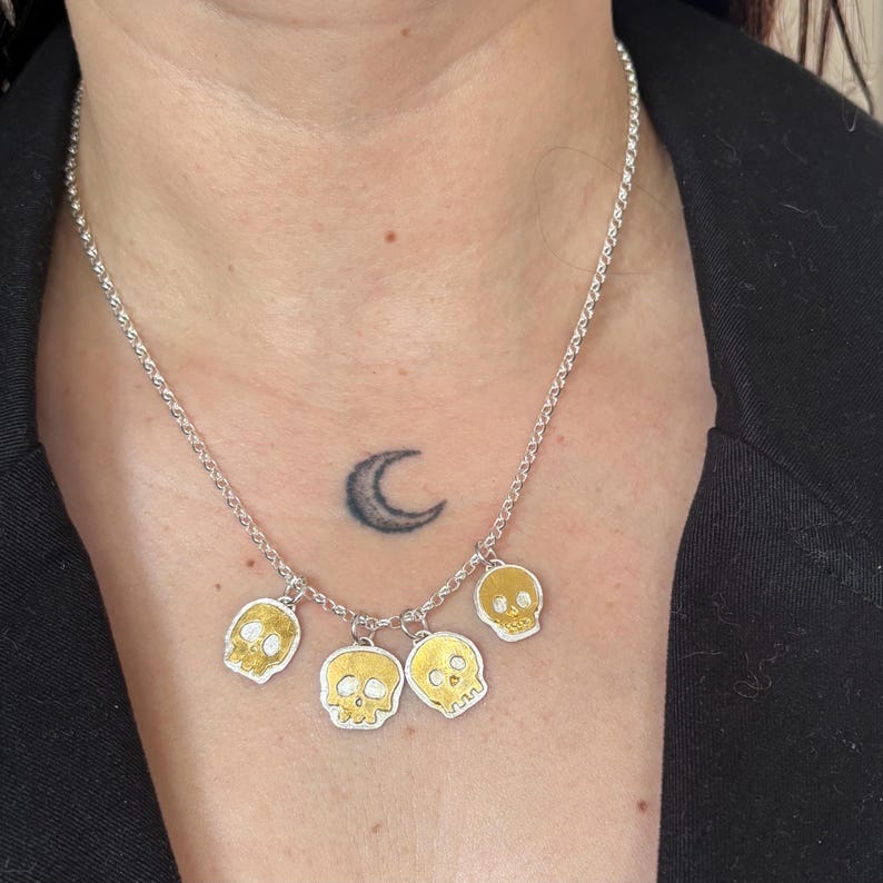 Op de afbeelding: Zilveren ketting met vijf goudkleurige schedelhangers. De schedels hebben gedetailleerde gezichtskenmerken en hangen aan een fijne ketting. De ketting wordt gedragen tegen een donkere achtergrond.