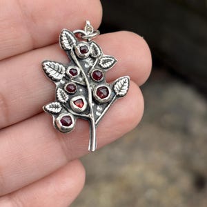 May include: A silver pendant shaped like a leafy branch with a textured surface. The branch features several small, faceted, deep red stones set into the metal. A small silver jump ring is attached to the top of the branch for hanging.