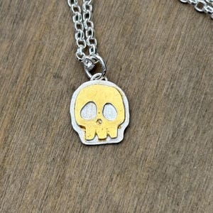 Op de afbeelding: Een zilveren ketting met een goudkleurige schedel hanger. De schedel hanger heeft een gouden midden en een zilveren rand. De hanger is een gestileerde weergave van een menselijke schedel.