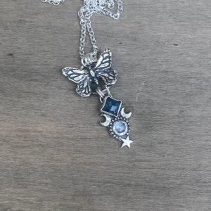 May include: A silver necklace with a butterfly charm, a blue gemstone, and a crescent moon charm. The necklace is on a wooden surface.