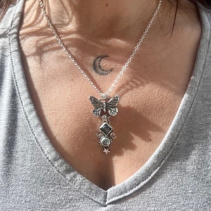 May include: A silver necklace with a butterfly charm and a moon and star pendant. The necklace is on a person's chest.