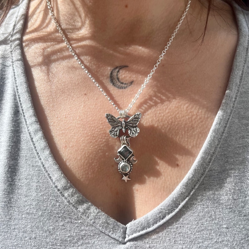 May include: A silver necklace with a butterfly charm and a moon and star pendant. The necklace is on a person's chest.
