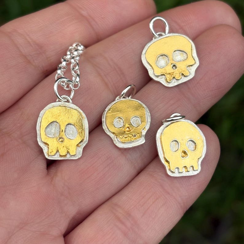 Op de afbeelding: Vier zilveren en gouden schedelhangers. E&eacute;n hanger zit aan een zilveren ketting. De schedels hebben een gouden midden en een zilveren rand. De hangers zijn in verschillende maten en ontwerpen, met zichtbare oog- en neusgaten.