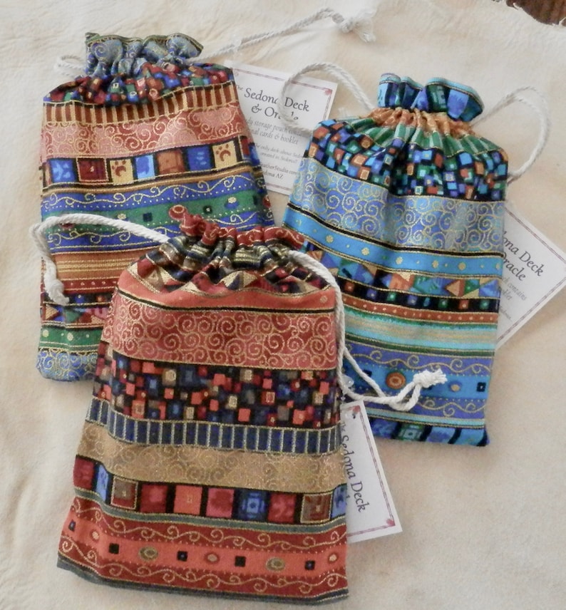 May include: Three colourful drawstring bags with gold trim and geometric patterns. Each bag has a tag that reads "Sedona Deck & Oracle".