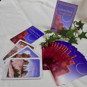 May include: A deck of oracle cards called "The Sedona Deck & Oracle Cards" with a purple and red design. The cards are fanned out on a white surface with a book and a green plant.