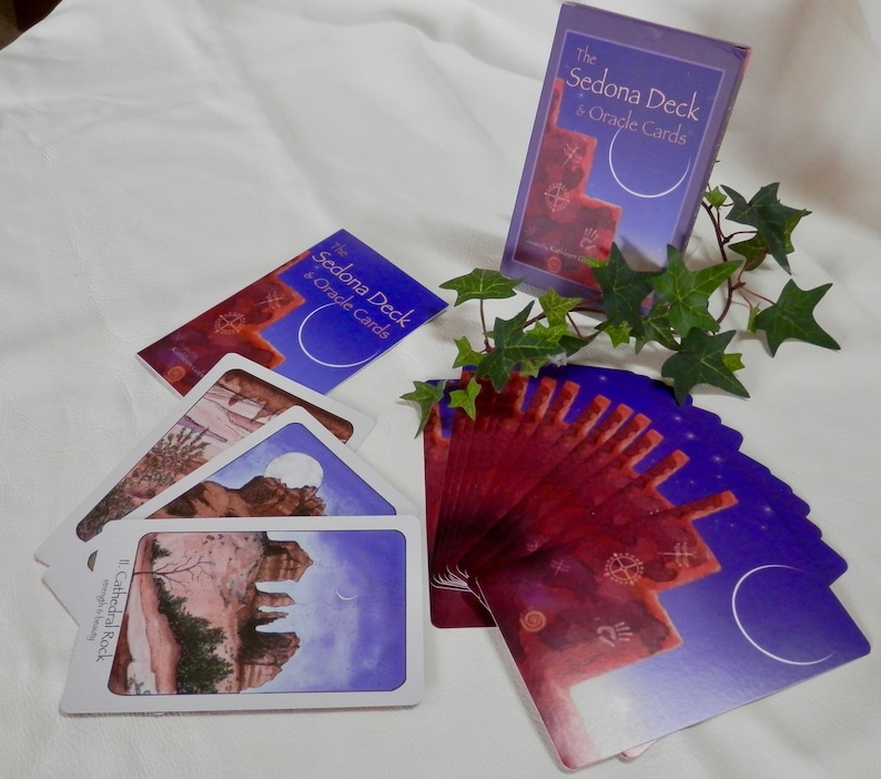 May include: A deck of oracle cards called "The Sedona Deck & Oracle Cards" with a purple and red design. The cards are fanned out on a white surface with a book and a green plant.
