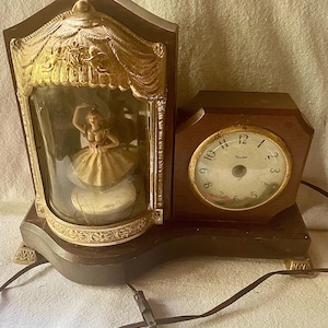 May include: A vintage wooden clock with a ballerina figurine inside a glass enclosure. The clock has a gold-colored decorative top and a round clock face with the word "United" on it. The clock is powered by a black cord.
