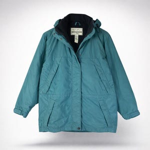 May include: Teal-colored hooded jacket with a black collar and lining. The jacket has a zipper closure, two front pockets, and a small logo tag on the chest. The jacket is designed for outdoor wear.