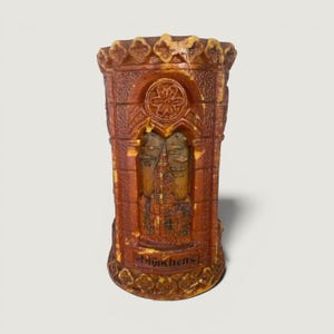 May include: A cylindrical brown wax candle featuring a relief sculpture of a gothic architectural structure. The design includes a circular emblem at the top and the word "Münchens" embossed at the base, with decorative scalloped edges.