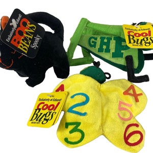 Puede incluir: Tres juguetes de peluche: un gato negro con una etiqueta "Boo Beans", un saltamontes verde con "GHP" en la espalda y una mariposa amarilla con los números 2, 3, 4, 5 y 6 en sus alas. Cada juguete tiene una etiqueta "Cool Bugs".