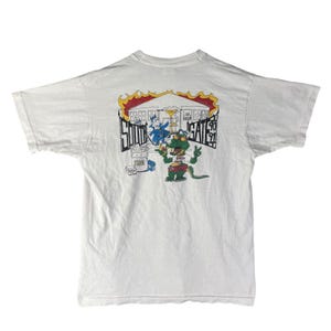 May include: White t-shirt featuring a colorful graphic on the back. The graphic includes cartoon characters, a building, and the text "SOUTH GATE 91". The design has a vintage, retro style.