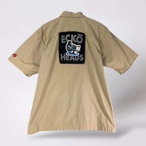 May include: Tan short-sleeved button-up shirt with a black patch on the back. The patch features the text "ECKO HEADS" and a graphic of a person on a skateboard. The shirt has a small red logo on the left sleeve.