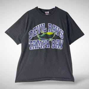 Pode incluir: T-shirt cinzento escuro com o texto "DEVIL RAYS TAMPA BAY" em branco e roxo. A camisa apresenta um gráfico de uma manta-raia em verde, preto e roxo. A t-shirt é de estilo vintage.