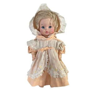 May include: A vintage doll with blonde hair and blue eyes, wearing a peach and cream-colored dress with a matching bonnet. The dress features lace trim and a bow at the waist. The doll's cheeks are rosy, and it has a classic, nostalgic appearance.