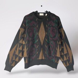 May include: A vintage crew neck sweater in dark gray with geometric and floral patterns. The design features brown diamond shapes, green and purple floral motifs, and brown triangle accents on the sleeves. The sweater is hanging on a black hanger.