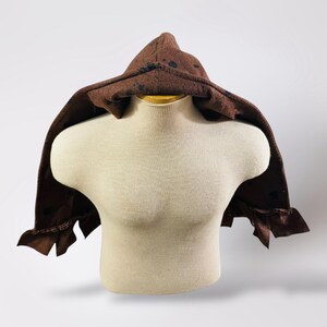 May include: A brown hooded cloak with black speckles, displayed on a mannequin. The cloak has a pointed hood and ruffled edges. The fabric appears to be a textured material, suitable for costume or cosplay.