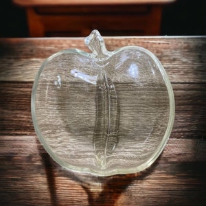 May include: A clear glass apple shaped dish with a divided center. The dish is perfect for serving snacks or dips.