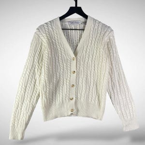 May include: Cream-colored cable knit cardigan sweater with a V-neck and button closure. The sweater is hanging on a black hanger. The brand name "Karen Scott" is visible on the label.
