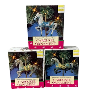 May include: Three boxes with hand-painted porcelain and brass carousel ornaments. Each box features a different colored horse with a blue, yellow, or green saddle. The boxes have a pink background with the text "Handpainted Porcelain and Brass... CAROUSEL ORNAMENT" on the front.