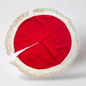May include: A round, red Christmas tree skirt with a cream-colored fringe border. The skirt has a slit for the tree trunk and is made of a soft, velvety material. The fringe adds a decorative touch to the holiday decor.