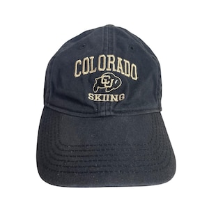 May include: A navy blue baseball cap with the word "COLORADO" embroidered in tan thread across the front. Below the word "COLORADO" is a tan embroidered graphic and the word "SKIING". The cap has a curved brim.