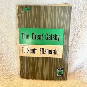 May include: A paperback copy of The Great Gatsby by F. Scott Fitzgerald. The cover features a dark olive green background with vertical gray lines, a central green horizontal band with black text, and a white band with the author's name in black.