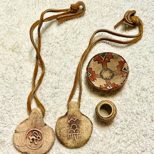 May include: Handmade Native American-style pendants and small bowl. Two pendants with leather cords feature embossed designs. A small, round bowl and a small, cylindrical container are also present. The bowl has a geometric pattern in red, brown, and beige.