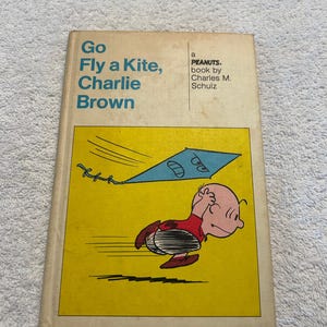 May include: A vintage copy of "Go Fly a Kite, Charlie Brown" by Charles M. Schulz. The book cover features Charlie Brown running with a kite against a yellow background. The title is in large teal letters.