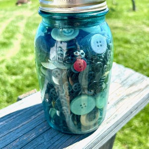 May include: A vintage-style blue glass mason jar with a silver metal screw-top lid, filled with a collection of assorted sewing buttons in various sizes, shapes, and colors, including a prominent single red button near the center.