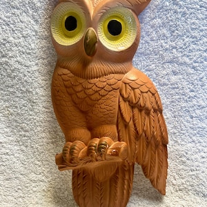 May include: A brown ceramic owl figurine with large yellow eyes and black pupils. The owl is perched on a branch and has detailed feather textures. The figurine is a decorative item.