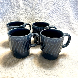 May include: Four matching blue ceramic mugs with a textured, ridged design. Each mug has a handle and a stacked base. The mugs are arranged on a white surface.