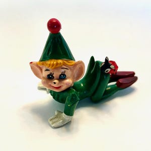May include: A vintage Christmas elf figurine. The elf is wearing a green outfit with a matching hat topped with a red ball. The elf has blonde hair, rosy cheeks, and blue eyes. The elf is lying down with its legs extended.
