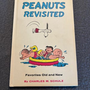 May include: A hardcover book titled Peanuts Revisited by Charles M. Schulz. The off-white cover features blue text and a colorful illustration of Peanuts characters in a yellow and red inflatable pool, with Snoopy diving from above.