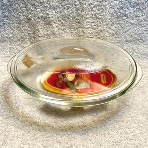 May include: A clear glass casserole dish with a matching lid. The dish has a rounded oval shape with handles on either side. The lid has a small handle in the center. The dish contains a red and yellow object.