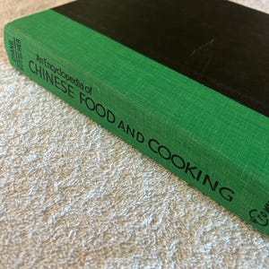 May include: A hardcover book titled "An Encyclopedia of Chinese Food and Cooking." The book has a black cover and a green spine with white text. The book is lying on a white, textured surface.