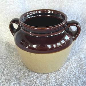 May include: A two-toned ceramic pot with a dark brown top and handles, and a tan base. The pot has a glossy finish and is likely used for cooking or storage. The pot is sitting on a white surface.