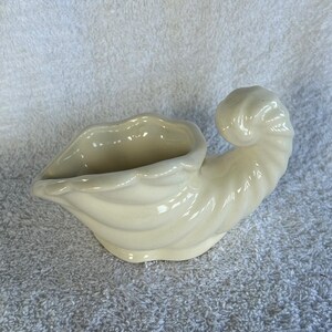May include: A cream-colored ceramic cornucopia vessel featuring a smooth, glossy finish and a spiraled, horn-shaped design. The opening is wide with a scalloped edge, tapering down to a curved, pointed tail that curls upward at the end.