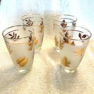 May include: Four frosted glass tumblers with gold leaf designs. Each glass has a gold rim and is decorated with gold and brown leaf patterns. The glasses are set on a white surface.