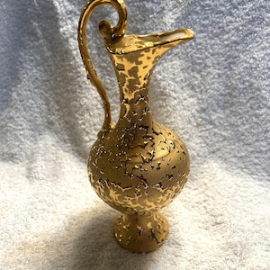 May include: A gold-colored decorative pitcher with a textured surface. The pitcher has a handle and a spout. The base is round and elevated. The overall design is ornate and vintage.