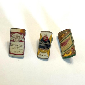May include: Three vintage beer can pins. The Budweiser pin is red and white with the brand name. The Stroh's pin is yellow, white, and black. The Miller High Life pin is gold and red. All three pins are rectangular.