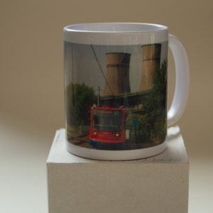 May include: White ceramic mug featuring a colourful image of a red tram travelling under a bridge with two large industrial towers in the background.