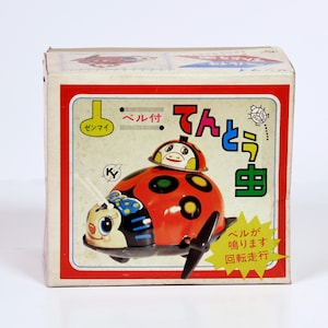 May include: A vintage toy ladybug in a box. The ladybug is red with black spots and a smiling face. The box has Japanese text and a red border.