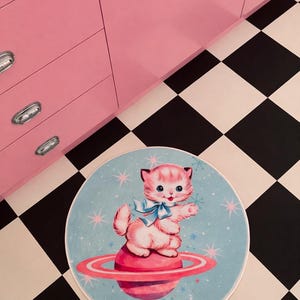 May include: A round rug featuring a pink kitten wearing a blue bow, sitting on a pink planet with a ring. The rug has a light blue background with star accents. The floor is black and white checkered.
