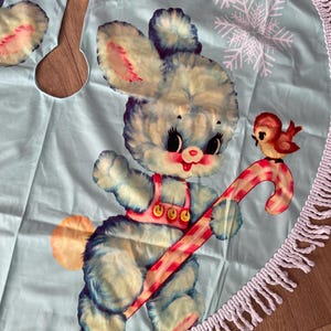 Pode incluir: Uma saia de &aacute;rvore de Natal azul claro com uma ilustra&ccedil;&atilde;o vintage de um coelhinho azul fofo segurando uma bengala doce. Um pequeno p&aacute;ssaro vermelho tamb&eacute;m est&aacute; presente. A saia tem uma borda de franjas brancas e detalhes de flocos de neve.