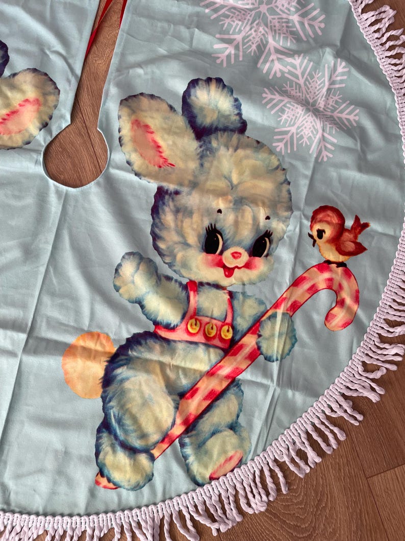 Pode incluir: Uma saia de &aacute;rvore de Natal azul claro com uma ilustra&ccedil;&atilde;o vintage de um coelhinho azul fofo segurando uma bengala doce. Um pequeno p&aacute;ssaro vermelho tamb&eacute;m est&aacute; presente. A saia tem uma borda de franjas brancas e detalhes de flocos de neve.