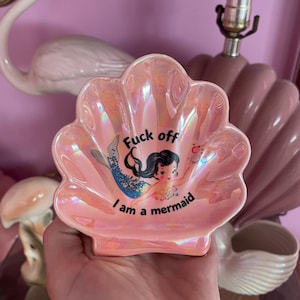 May include: A shell-shaped trinket dish with an iridescent pink finish. The centre features a graphic of a cartoon mermaid with black hair and a blue tail, accompanied by the black text "Fuck off I am a mermaid" printed in a curved layout.